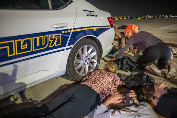 Mitglieder der Delegation von Bundeskanzler Olaf Scholz liegen auf dem Flughafen von Tel Aviv hinter einem Polizeiwagen auf dem Boden. Foto: picture alliance/dpa Pool/Michael Kappeler